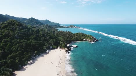 aerial-view-of-the-beach-at-Tayrona-Natural-Park-in-Santa-Marta,-featuring-crystal-blue-ocean-waters-and-lush-green-trees-lining-the-shore