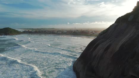 Wellen,-Die-Gegen-Die-Schroffen-Klippen-Schlagen,-Mit-Dem-Strand-Im-Hintergrund,-Beleuchtet-Vom-Warmen-Schein-Der-Sonne