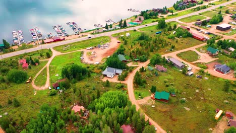 Cars-Driving-on-Road-Between-Beautiful-Clear-Blue-Lake-Water-and-Forest-with-Houses-and-Boat-Docks