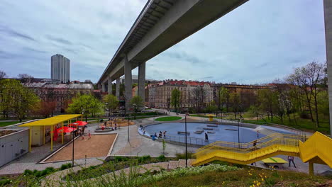 La-Carretera-Del-Puente-Cruza-Sobre-La-Infraestructura-Del-Parque-Urbano-De-La-Ciudad-De-Praga,-República-Checa,-Con-Gente-Relajándose-Y-Pasando-El-Rato-En-El-área-Urbana,-Horizonte-Nublado