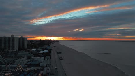 An-aerial-view-of-the-boardwalk-and-beach-at-Coney-Island,-NY-during-a-cloudy-but-colorful-sunrise