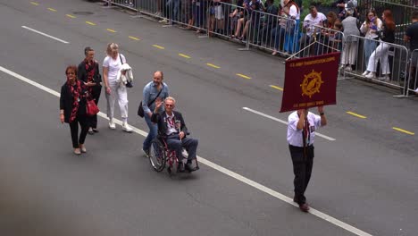 Senior-veteran-from-the-US-Army-Transportation-Corps,-in-a-wheelchair-is-being-pushed-down-the-street-by-his-family,-waving-at-the-cheering-crowds-during-the-Anzac-Day-parade-in-Brisbane-city