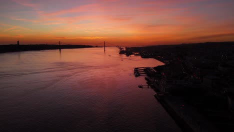 Last-light-fire-sunset-dusk-glow-spreads-pink-hazey-clouds-over-Tagus-river-in-Lisbon-Portugal,-silhouette-of-bridge