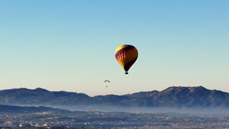 Stationäre-Drohnenansicht-Eines-Heißluftballons-Und-Eines-Gleitschirms,-Die-Von-Rechts-Nach-Links-über-Temecula-Weingütern-Mit-Klarem-Himmel-Und-Tief-Hängendem-Nebel-Und-Hügeln-Im-Hintergrund-Vorbeisegeln