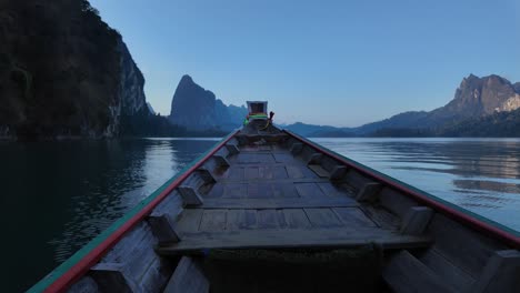 Timelapse-clip-showing-front-of-wooden-boat-moving-over-calm-lake-in-Thai-National-park,-with-steep-rocky-mountains-and-native-rainforest