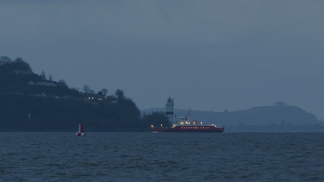 Distant-red-ferry-boat-on-the-water-coming-into-port-and-passing-lighthouse-on-land-on-cloudy-day