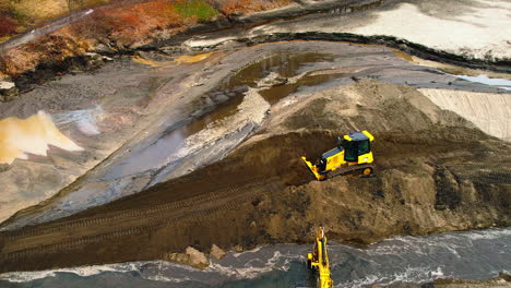 Tiro-De-Seguimiento-Aéreo-Bulldozer-Amarillo-Empujando-La-Tierra-En-Un-Nuevo-Camino-De-Tierra-En-La-Orilla-De-Un-Pequeño-Río