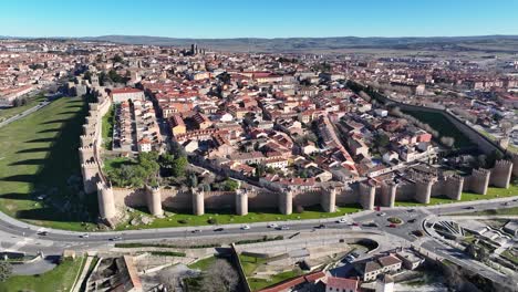 Flug-Mit-Einer-Kameradrehung,-Die-Mit-Einem-Blauen-Himmel-Die-Stadt-Avila-Visualisiert,-Ein-UNESCO-Weltkulturerbe-Mit-Seiner-Steinmauer-Und-Seinen-Türmen-Und-Einer-Umgehungsstraße-Mit-Fahrzeugen,-Die-In-Spanien-Zirkulieren