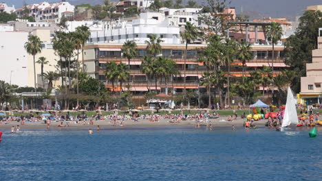 Los-Christianos-Beach-in-Tenerife---with-sunbathers,-swimmers,-and-kayakers-against-a-backdrop-of-palm-trees-and-buildings