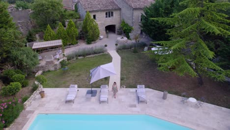 A-young-owner-leaves-her-stone-farmhouse-to-go-to-her-swimming-pool,-the-shot-accompanies-her-as-she-moves-backwards,-revealing-the-village-and-nature-and-its-fir-trees