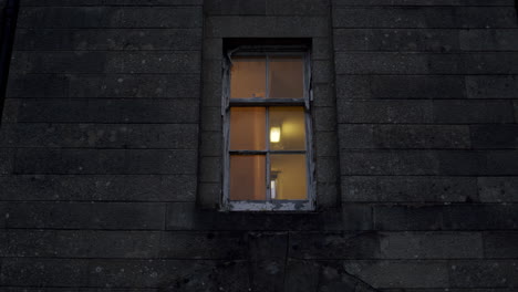 Wooden-window-with-the-light-shining-in-apartment-on-a-grey-brick-wall-of-a-building,-handheld-urban-location