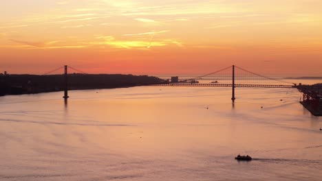 Classic-city-scene-of-tug-boats-and-cars-commuting-across-suspension-bridge-at-mouth-of-waterway-at-dusk-with-orange-sunset