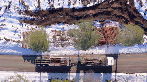 Truck-loaded-with-harvest-tree-logs-pulling-away-in-winter,-top-down-aerial