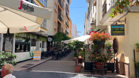 Walking-through-Estepona-old-town-street-where-people-sit-in-restaurants-outside,-sunny-day-in-Andalusia-Spain,-vacation-holiday-destination,-4K-shot