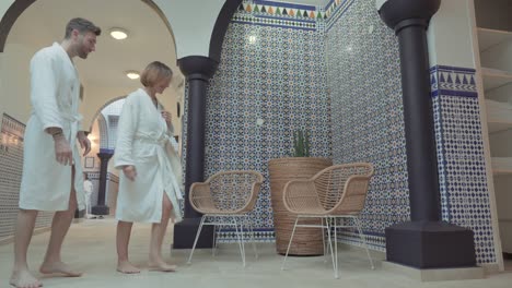 A-young-couple-in-white-bathrobes-walks-down-the-corridor-of-a-spa-to-sit-on-wicker-chairs