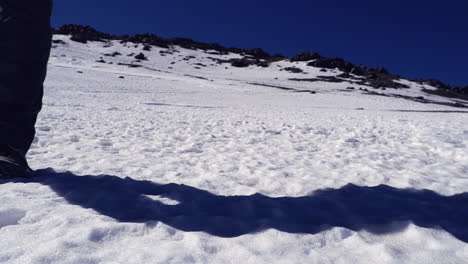 Low-angle-hiker-walks-to-camera-from-snowy-mountain-ridge,-blue-sky