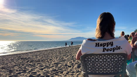 Mujer-Sentada,-Hablando-Y-Disfrutando-De-La-Vida-En-Un-Restaurante-De-Playa-En-Marbella,-Mujer-Vistiendo-Un-Suéter-Con-Palabras-De-Gente-Feliz,-Día-Soleado-De-Verano-En-España,-Toma-De-4k