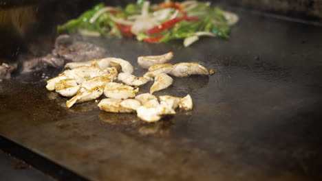 Spatula-spread-out-steaming-sizzling-beef-fajita-strips-on-flat-top-grill,-chicken-and-beef-fajitas-with-peppers-and-onions-cooking-in-commercial-restaurant-kitchen-griddle,-slow-motion-close-up-4K