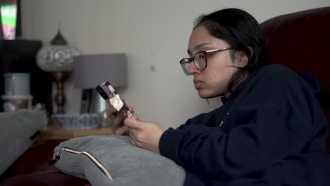 Young-British-South-Asian-Female-enjoying-ice-cream-while-lounging-on-a-sofa-in-a-cozy-home-environment,-casually-glancing-at-the-display-of-a-mobile-phone-screen