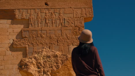 Mujer-Turista-Asiática-Visitando-Egipto-Parada-Frente-A-Jeroglíficos-Egipcios-Tallados-En-Una-Pared-Roja-En-El-Antiguo-Egipto,-Lengua-Perdida-Egipcia-En-El-Desierto-De-Oasis-En-El-Norte-De-áfrica