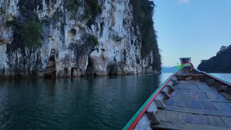 Side-Profile-of-Wood-Boat-Cruising-Through-Ocean-Water-By-Large-Vegetated-Rocks,-Thailand