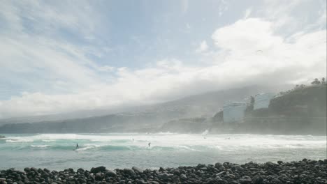 Slowmotion-group-of-surfers-surfing-on-ocean-waves,-red-flag-on-pebble-beach,-Playa-Martiánez,-Puerto-de-la-Cruz,-Tenerife,-Canary-Islands-in-spring