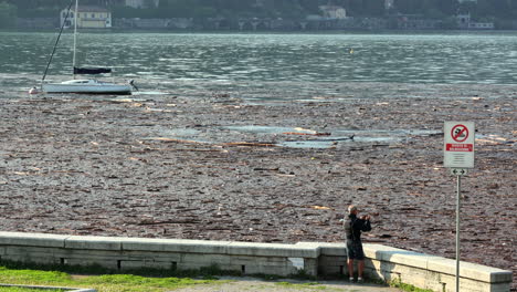 Como,-Italy---july-29-2021---a-man-taking-photos-of-the-lake-covered-in-timber-and-debris-after-heavy-rains-that-caused-severe-damage-in-the-area