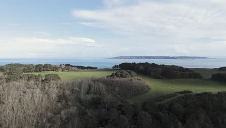 Flight-over-Herm-Island,-Channel-Islands-view-towards-Sark-and-panning-over-the-Island-with-trees-and-fields-to-Shell-Beach-at-low-tide-with-clear-calm-sea