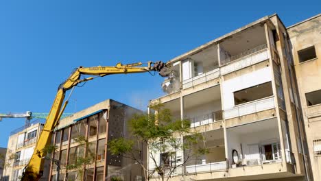 Tall-yellow-excavator-crane-with-claw-attachment-demolishes-upper-level-roof-of-apartment-building