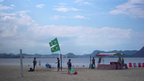 A-perspective-of-Copacabana-Beach-in-Rio-de-Janeiro,-Brazil,-showcasing-locals-relaxing-amidst-two-Brazilian-flags-against-the-backdrop-of-expansive-blue-skies