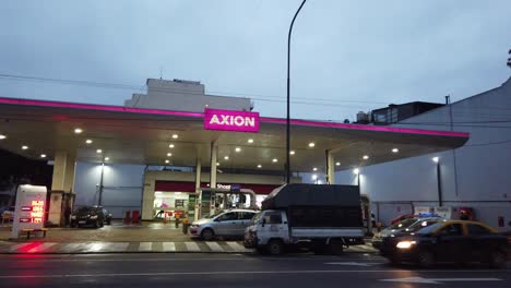 Axion-pink-magenta-gas-petrol-oil-station-at-night-panoramic-avenue-cars-drive-fast-at-buenos-aires-cityscape,-nighttime-lights-city-neighborhood,-Flores,-Gaona