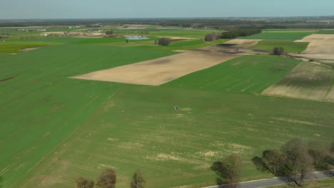 Aerial-eveals-a-patchwork-of-farmland,-where-a-solitary-tractor-methodically-sprays-crops,-etching-lines-of-progress-across-the-verdant-landscape