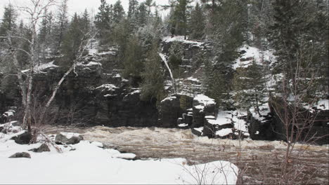 River-flows-by-a-snow-covered-rocky-cliff-covered-in-pines,-with-snowflakes-softly-drifting-down