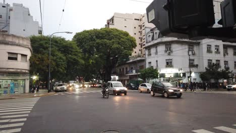 Car-traffic-drives-by-directorio-avenue-in-latin-american-dusk-skyline-people-wait-for-crossing-trees-architecture