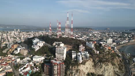 Aerial-Drone-Fly-Resorts-at-Cliff-next-to-Pacific-Ocean-Beach-in-Mazatlan-Mexico-Waves-crushing-coastal-town,-sea-and-skyline-along-5G-communication-towers