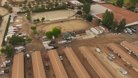 Drone-Shot-Over-Livery-Stables-at-Burbank-Equestrian-Center-in-Daytime,-Horses-Running-Around-in-Training-Pen-with-Trailers-and-Trucks-Moving-Around-the-Lot