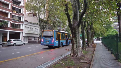 Blue-autobus-stops-at-park-urban-green-space,-wet-humid-avenue-in-latin-american-city,-buildings-asphalted-road,-buenos-aires-city-argentina,-caballito-neighborhood