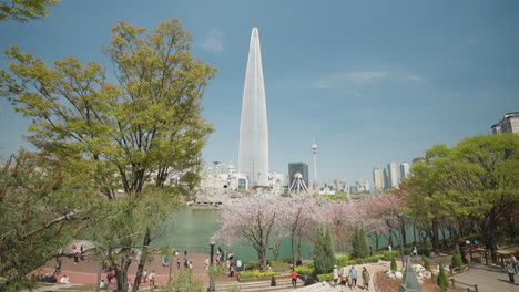 Lottle-World-Tower-Against-Blue-Sky-in-Spring,-Seoul-Citizens-Enjoy-Cherry-Blossom-Walking-Around-Seokchon-Lake-in-Songpa-Naru-Park