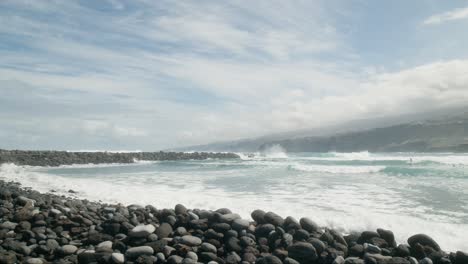 Slowmotion-ocean-waves-crushing-on-pebble-beach,-group-of-surfers,-Playa-Martiánez,-Puerto-de-la-Cruz,-Canary-Islands-in-spring