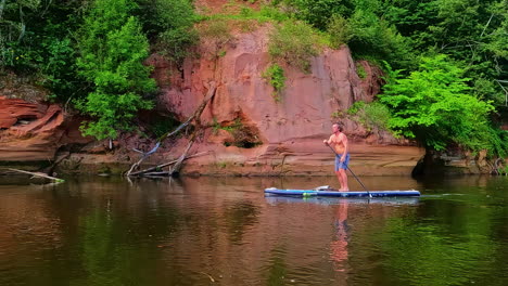 Man-paddleboarding-along-the-Erglu-Cliffs-of-the-Gauja-River-and-national-park,-Latvia