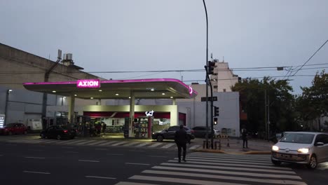 Ubicación-De-La-Gasolinera-Por-La-Noche,-La-Gente-Cruza-La-Avenida-En-La-Ciudad-Latinoamericana,-Los-Automóviles-Circulan-Por-Axion-Pink-Display-Oil-Landmark