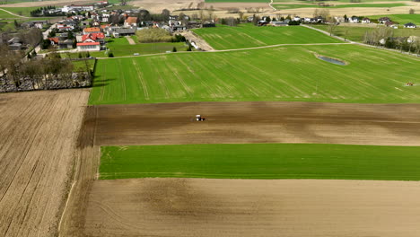 Aerial-birds-eye-of-plowing-tractor-on-farm-field-near-small-village-at-sunny-day-in-spring-season