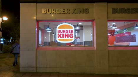 Los-Peatones-Pasan-Por-La-Cadena-Estadounidense-De-Restaurantes-De-Comida-Rápida-De-Hamburguesas,-Burger-King-Y-El-Cartel-Con-El-Logotipo-Durante-La-Noche.