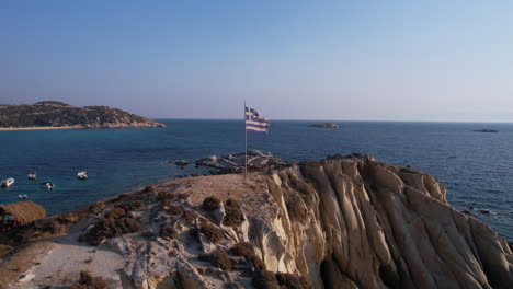 Bandera-Nacional-De-Grecia-Ondeando-En-Un-Poste-En-Un-Acantilado-Sobre-La-Playa-Y-El-Mar-Egeo,-Disparo-De-Drones