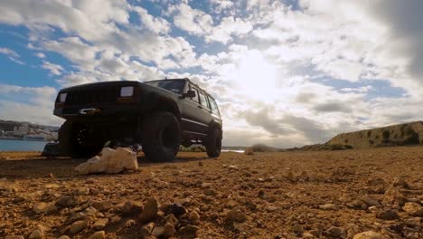Description:-A-Jeep-Cherokee-stands-on-rocky-ground-as-a-time-lapse-captures-swiftly-moving-clouds-above