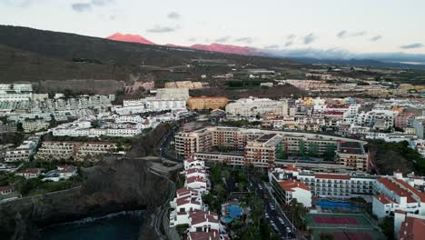 The-drone-captures-Los-Gigantes-town-with-its-picturesque-coastal-charm,-while-in-the-distance,-the-majestic-red-hued-Teide-volcano-stands-as-an-iconic-landmark-on-Tenerife-island