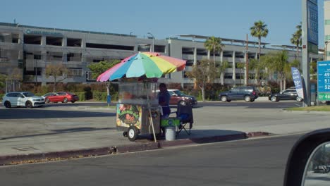 Street-vendor-selling-snacks-under-colorful-umbrella-in-sunny-Los-Angeles,-cars-passing-by,-urban-setting