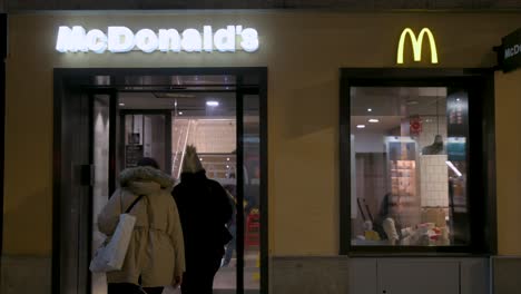 Por-La-Noche,-Los-Clientes-Entran-En-Un-McDonald&#39;s,-Una-Cadena-De-Comida-Rápida-Estadounidense,-Mientras-Una-Niña-Disfruta-De-Una-Comida-En-El-Interior,-Visible-A-Través-De-La-Ventana.