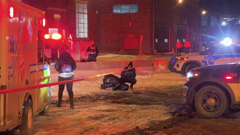 Emergency-services-tending-to-a-crying-person-after-a-fatal-shooting-at-night-with-police-tape,-ambulances-and-officers-at-a-crime-scene-in-Montreal