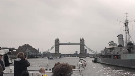 View-from-a-ferryboat-on-a-Thames-River-cruise-in-London,-passing-HMS-Belfast-with-Tower-Bridge-in-the-backdrop